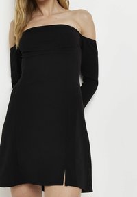 Černé šaty bez ramínek s dlouhými rukávy, přiléhavým korzetem a mírným bočním rozparkem, vyrobené z hladké, elastické látky.