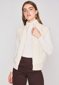 Canotta senza maniche color crema con una parte anteriore morbida e texture e bordi in maglia a coste; presenta tasche laterali e una cerniera frontale. Indossata sopra un dolcevita bianco.