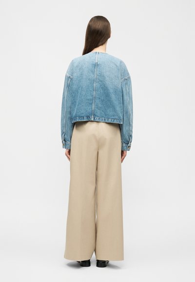 Άτομο που στέκεται με την πλάτη γυρισμένη, φορώντας ένα ανοιχτό γαλάζιο τζιν μπουφάν, μπεζ παντελόνι φαρδιάς γραμμής και μαύρα παπούτσια, σε λευκό φόντο.