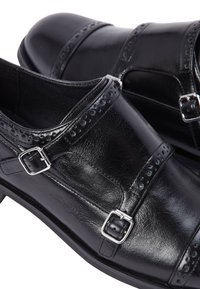 Chaussures en cuir noir avec une finition lisse, des motifs perforés ornementaux, et deux boucles en argent sur le dessus et la sangle de la cheville.