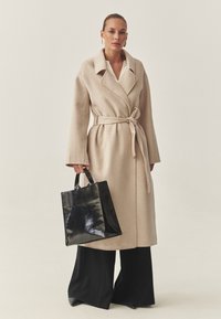 Bež volnen plašč z pasom v pasu, velikimi ovratniki in širokimi rokavi. Črna torba iz lakastega usnja. Črne široke hlače.