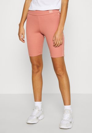Pantaloni bici rosa a coste con vita alta. Indossati con sneakers bianche e calzini alla caviglia. Il design presenta un logo sulla cintura.