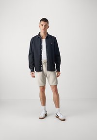 Denimowa koszula w ciemnoniebieskim kolorze z zapięciem na guziki, zestawiona z jasnobeżowymi szortami. Model nosi białą koszulkę bez rękawów oraz sneakersy z brązowymi akcentami.