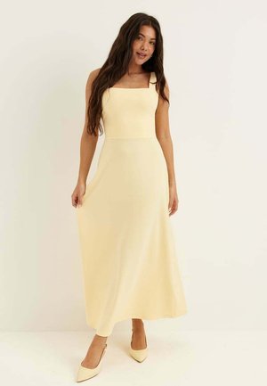 Femme aux longs cheveux foncés portant une robe maxi jaune pâle sans manches et des escarpins pointus assortis, debout devant un fond uni.