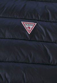 Navy puffer jacket with a quilted texture, featuring a triangular white and red "GUESS" logo badge and outlined accents.