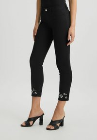 Jeans din bumbac negru cu tiv brodat, design ajustat, talie la nivel mediu și stil cu cinci buzunare, asortați cu pantofi cu toc cu bretele încrucișate negre.