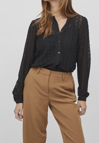 Blouse en dentelle noire avec un design à boutons sur le devant et des manches longues, associée à un pantalon taille haute beige. Tissu texturé avec des détails soignés.