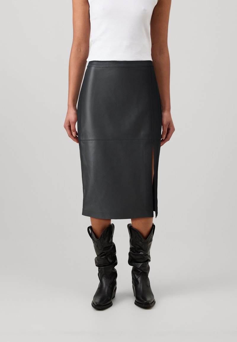 Femme portant une jupe midi en cuir noir avec une fente latérale, associée à des bottes de cowboy noires décontractées et un haut blanc sans manches.