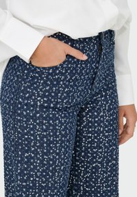 Pantalons larges navy ornés de sequins avec une finition en tissage texturé et des poches latérales. Gros plan sur une main reposant dans la poche.