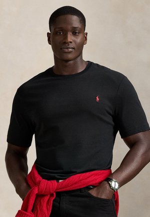 Μαύρο κοντομάνικο μπλουζάκι με κόκκινο κεντητό λογότυπο. Φοριέται με κόκκινο πλεκτό πουλόβερ δεμένο γύρω από τη μέση. Ομαλή υφή.