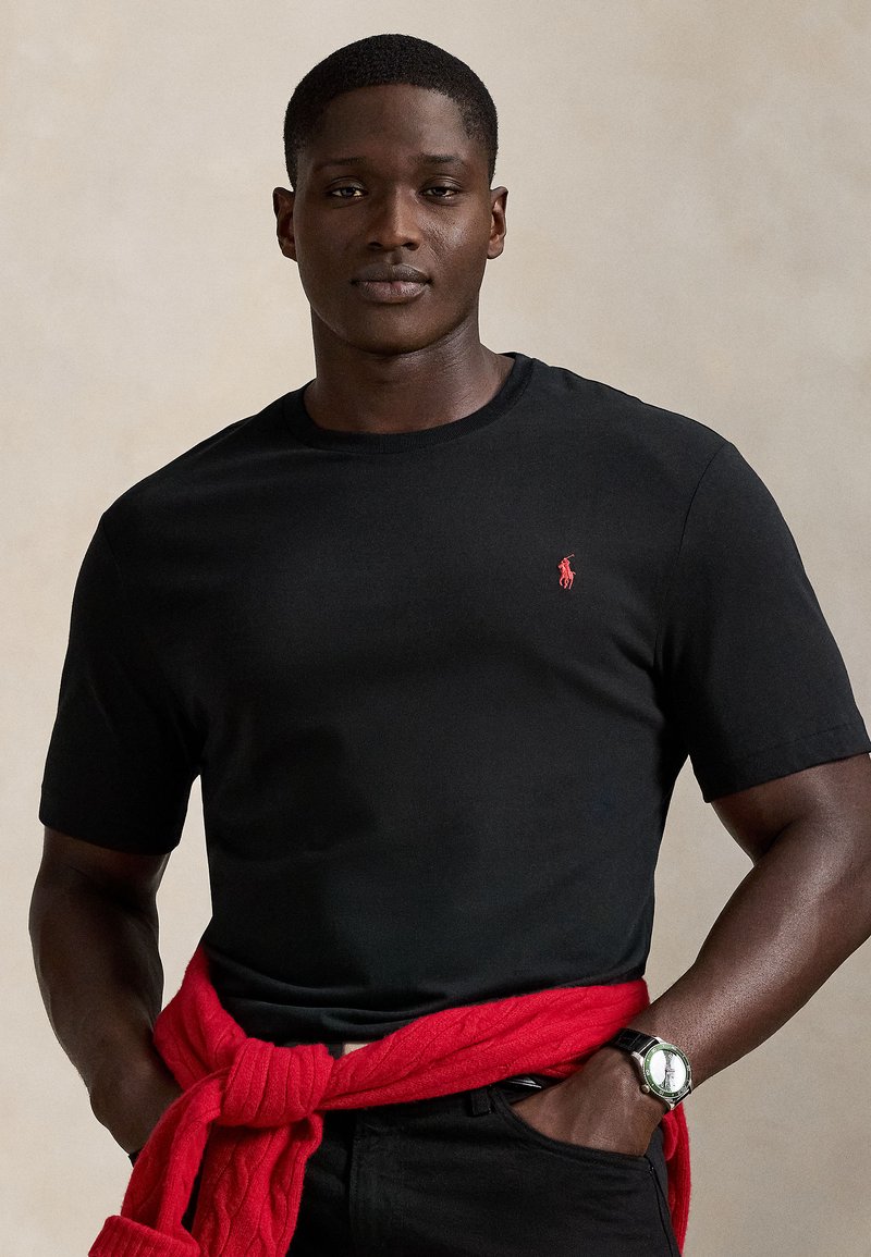 Černé tričko s krátkým rukávem a červeným vyšívaným logem. Nošeno s červeným pleteným svetrem uvázaným v pase. Hladká textura.