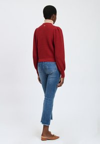 Rode gebreide trui met pofmouwen, crèmekleurige kraag en details op de rug. Gecombineerd met blauwe distressed jeans en lichtbruine platte schoenen.