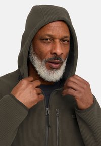 Olijfgroene geribde hoodie met een ritssluiting aan de voorkant, een capuchon met trekkoorden en twee zichtbare ritsockets. Zachte textuur met een warme voering.