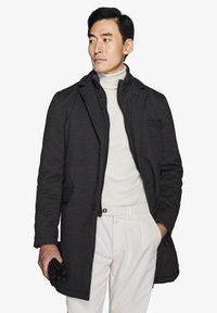 Sort uldfrakke med klapkrave, lagdelt over en lys turtleneck, parret med hvide bukser og sorte læderhandsker.