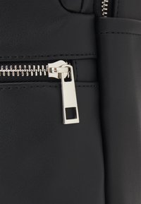 Sac en cuir noir avec une fermeture éclair argentée munie d'une tirette rectangulaire, une surface texturée et des accents cousus.