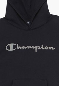 Svart hoodie med framfickor, prydd med en framträdande vit "Champion"-logotyp i skriptstil, tillverkad av mjukt bomullsmaterial.