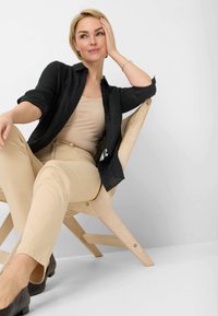 Chemise noire à boutons, débardeur beige et pantalon. Assis sur une chaise en bois claire au design incliné. Peau lisse, cheveux blonds courts. Fond neutre.