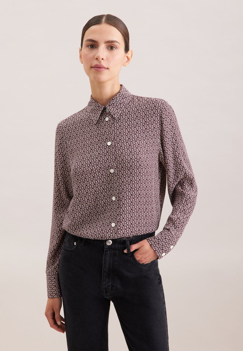 Chemise à manches longues bordeaux avec un motif géométrique, col boutonné et boutons sur le devant ; accompagnée de jeans noirs foncés.
