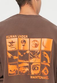 Brun sweatshirt med orange grafiktryk på ryggen, der viser ni firkanter med forskellige visuelle elementer og teksten "HUMAN UNDER MAINTENANCE."