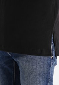 Fekete pamut póló, amelynek az alján oldalsó hasítékok találhatók, kék denim farmernadrággal párosítva, amely textúrált felülettel és látható varrással rendelkezik.