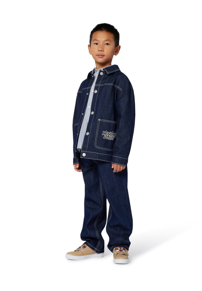 Ensemble de veste et de pantalon en denim, bleu foncé avec des coutures blanches. Comprend une poche frontale et une chemise rayée en dessous. Baskets beiges avec des détails brodés.