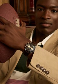 Montre chronographe avec un cadran noir, des accents dorés et un bracelet en cuir marron. Un ballon de football marron et blanc tenu dans une main, contre une veste beige.