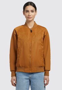 Giubbotto bomber bronzo con ricamo floreale sul petto, colletto e polsini a coste, e tasche laterali, abbinato a jeans azzurri chiari.