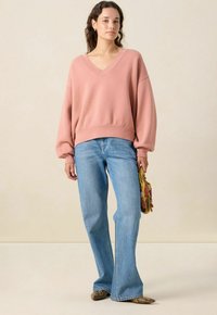 Sweatshirt rose surdimensionné avec un col en V, associé à un jean bleu évasé et un sac jaune à motif. Des chaussures plates en cuir de serpent complètent le look.