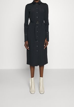 Žena v černých midi šatech s dlouhým rukávem a knoflíky, se zavázaným páskem v pase a bílými kotníkovými botami, stojící před jednoduchým pozadím.