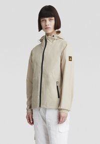 Giacca leggera beige con cappuccio, dotata di una cerniera nera, polsini elasticizzati e una toppa con logo sulla spalla. Texture liscia e design minimale.