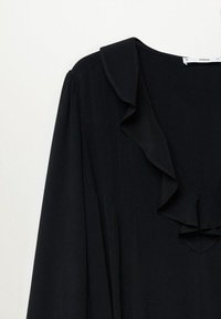 Blouse noire à manches longues avec un col en V à volants et une subtile texture verticale sur le tissu, présentée sur un fond blanc.