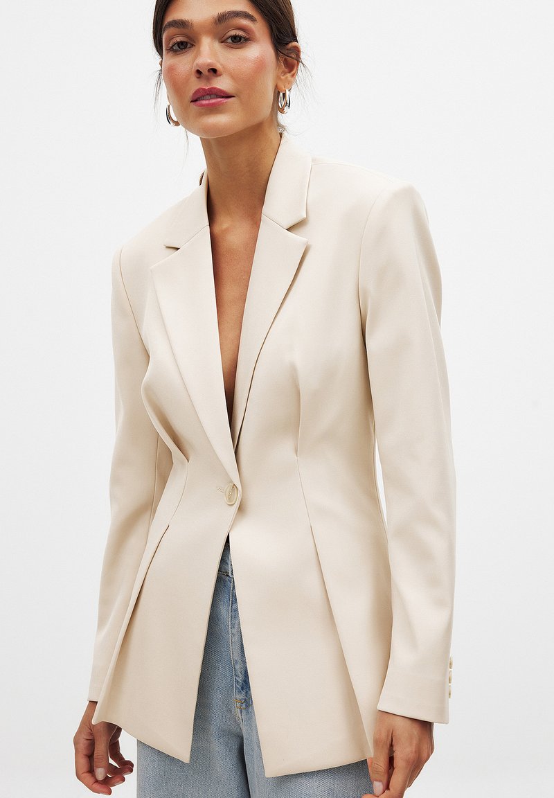 Blazer beige ajusté avec revers en pointe, fermeture à un bouton et épaules structurées. Le tissu lisse et l'ourlet évasé rehaussent le design.