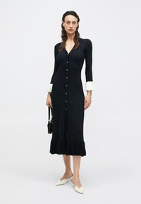 Czarna prążkowana sukienka midi z dekoltem w serek, zapięciem na guziki z przodu, rozkloszowanymi mankietami i falbaną u dołu. Noszona z białymi butami w szpic.