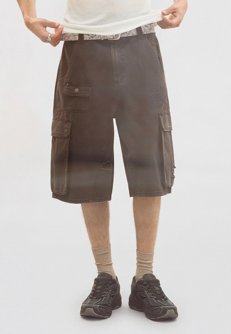 Homme portant un short cargo marron foncé, des chaussettes beige et des baskets noires, tenant l'ourlet d'une chemise blanche.