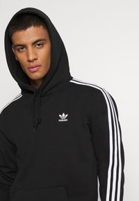 Czarna bluza z kapturem z białymi trzema paskami na rękawach i minimalistycznym białym logo Adidas. Posiada sznurki do regulacji kaptura oraz kieszeń kangurką.