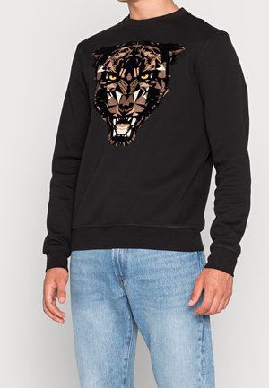 Mann trägt schwarzen Sweatshirt mit geometrischem Design eines fauchenden Pantherkopfes und hellblauer Jeans vor einfarbigem Hintergrund.
