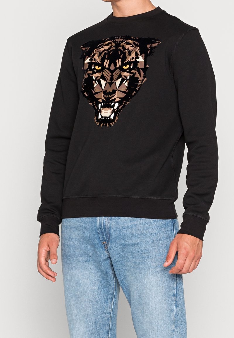 Homme portant un sweat-shirt noir avec un motif géométrique de tête de panthère rugissante et un jean bleu clair sur fond uni.