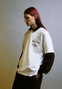T-shirt bianco oversize con colletto nero e maniche lunghe, dotato di una stampa grafica grigia. Tessuto in cotone morbido con vestibilità rilassata.