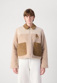 Veste en polaire beige avec un col en velours côtelé marron et deux poches avant. Présente une fermeture éclair dorée et un petit logo sur la poitrine.