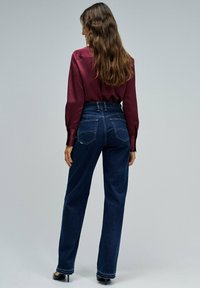 Jeans de mezclilla oscuros de tiro alto y pierna ancha con cinco bolsillos y un ligero efecto de desgastado. Se lleva una blusa de satén color burdeos encima.