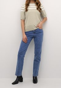 Kvinde iført beige kortærmet stribet striktrøje, blå lige ben jeans og sorte hælede ankelstøvler, stående mod en ensfarvet væg.