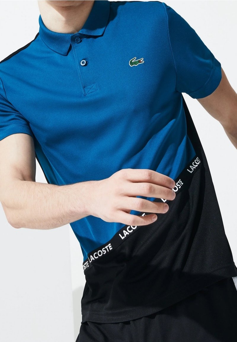 Homme portant un polo Lacoste bleu et noir avec le logo crocodile et le nom de la marque imprimés sur la partie noire.