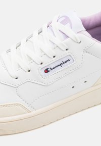 Sneakers bianchi con tomaia in pelle testurizzata, punta rotonda e suola in gomma off-white a contrasto. Presentano un'etichetta con logo e lacci bianchi.