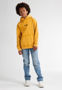 Felpa gialla con tasca frontale e logo, abbinata a jeans in denim azzurri e sneakers bianche. La felpa è dotata di cordino e polsini a coste.