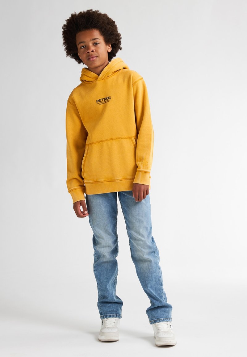 Felpa gialla con tasca frontale e logo, abbinata a jeans in denim azzurri e sneakers bianche. La felpa è dotata di cordino e polsini a coste.