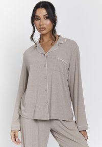Conjunto de pijama beige que incluye una camiseta de manga larga con collarete, botones en la parte delantera, bolsillo en el pecho y ribete blanco; textura suave con canalé.
