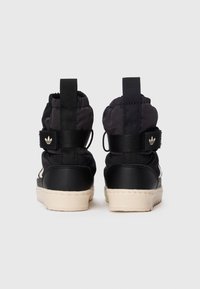 Une paire de baskets montantes noires avec une tige en tissu rembourré, des sangles noires, un logo à l'arrière et des semelles en caoutchouc beige, vues de l'arrière.
