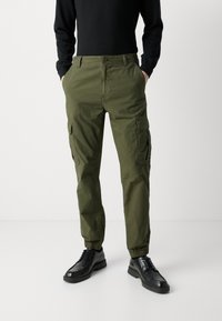 Calças cargo verde oliva com punhos elásticos, dois bolsos laterais e dois bolsos cargo. Usadas com sapatos pretos e uma camisa de manga longa preta.