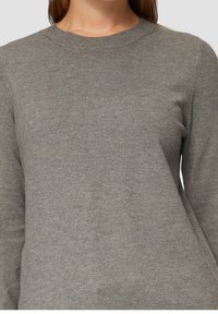 Kvinna klädd i en enkel grå långärmad stickad tröja med rund halsringning, sedd från halsen till midjan.