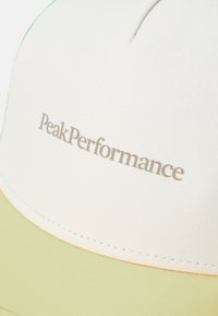 Svetlobéžová čiapka s zelenými akcentmi, mäkká tkanina, zakrivený šilt a logo "PeakPerformance" vyšívané svetlošedou niťou.